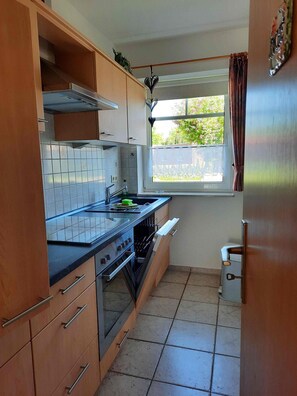 A compact kitchen with wooden cabinets, a tiled backsplash, a window with curtains, and a stainless steel range hood.