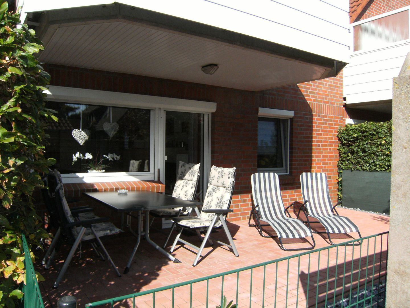 A patio with a table and two striped chairs, a window with decorative items, and a brick building in the background.