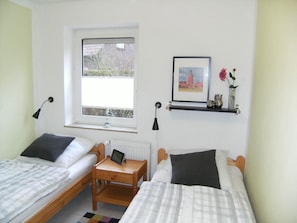 A room with two single beds, a small table with a tablet, a window with a view of houses, a framed picture on the wall, and a shelf with decorative items. - Apartment Jonathan Whg. 5 - Apartment Tulpenweg 4 Jonathan Whg. 5 (Neuharlingersiel)