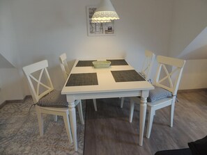 Uma área de refeições com mesa branca, quatro cadeiras, um cesto de vime e um abajur rendado.