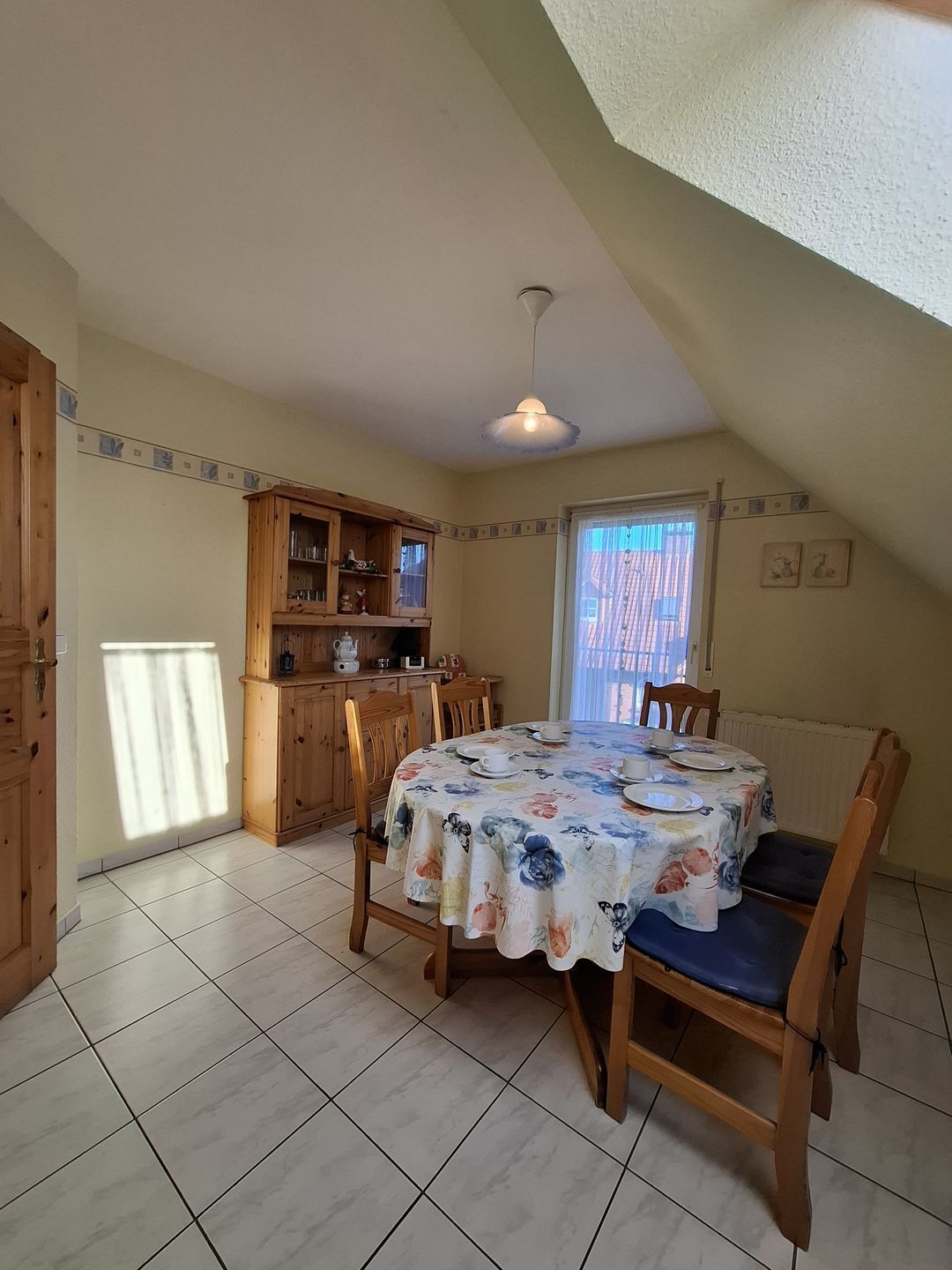 Ein Essbereich mit einem runden Tisch, der mit einer blumigen Tischdecke bedeckt ist, Holzstühle und eine Küchenzeile mit Holzschränken und einem Fenster.