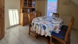 A dining area with a round table covered in a floral tablecloth, wooden chairs, and a kitchen area with wooden cabinets and a window. - House Klabautermann - House Klabautermann Blockshausen 1 Apartment 2 (Neuharlingersiel)