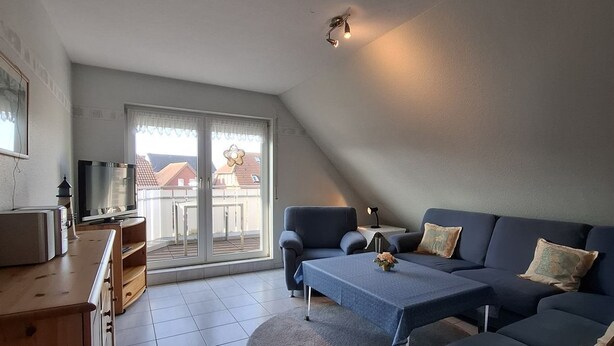 A modern living room with a sofa, a coffee table, and a TV. There is a balcony with a view of buildings. - House Klabautermann - House Klabautermann Blockshausen 1 Apartment 2 (Neuharlingersiel)