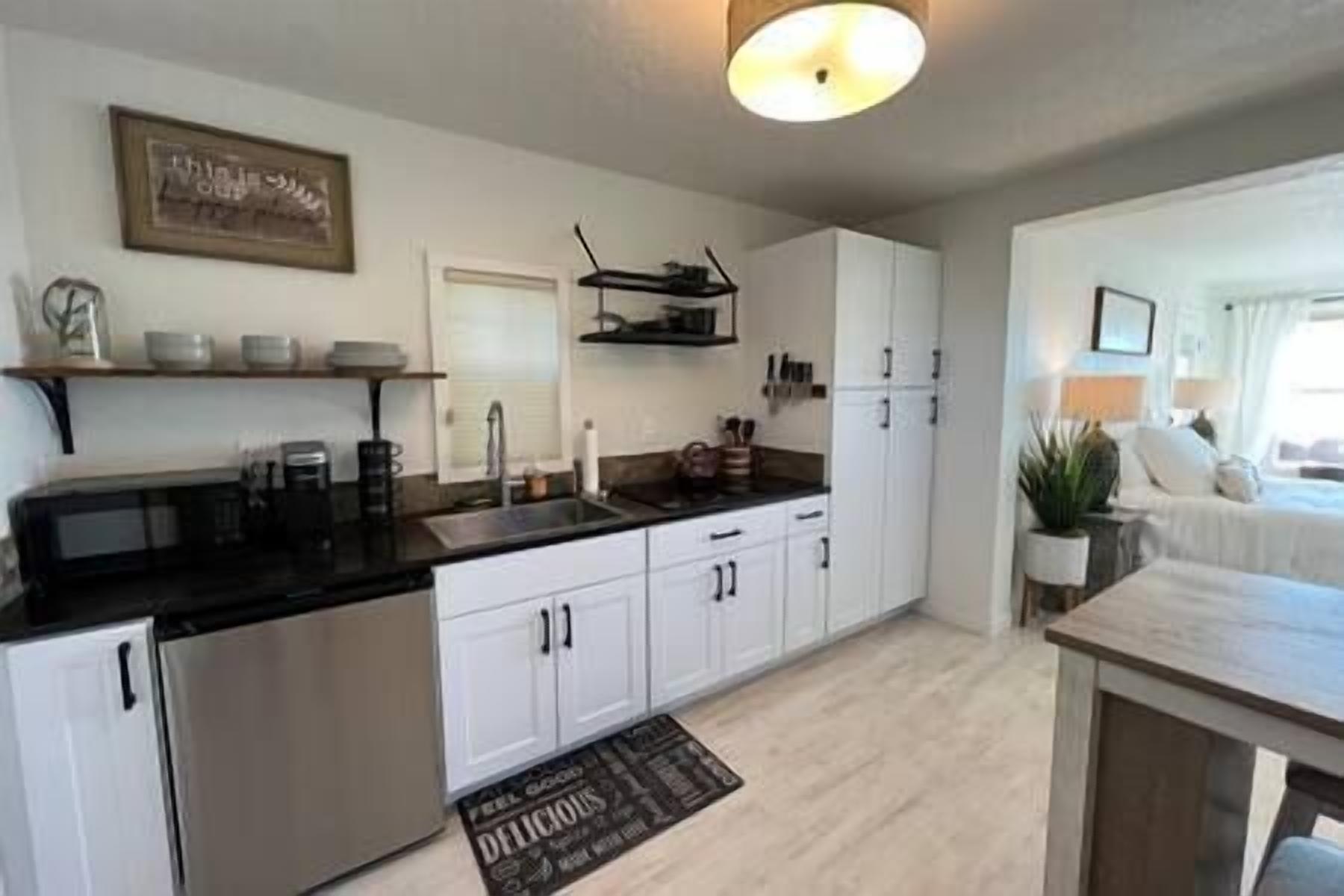 Une cuisine moderne avec des armoires blanches, un réfrigérateur en acier inoxydable et un comptoir noir. On y trouve un petit îlot central doté d’un évier et d’une étagère suspendue au-dessus.