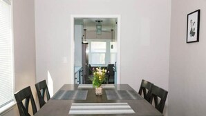 Een eetkamer met een donker houten tafel, zes stoelen en een bloevas. Er hangt een lijstje aan de muur en aan de linkerzijde is een raam.