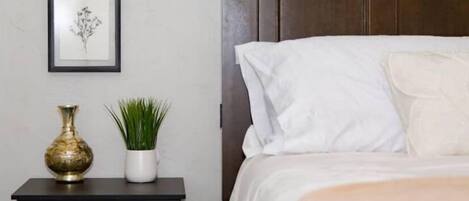A neatly made bed with white bedding, a dark nightstand with a brass vase and a potted plant, and a framed picture on the wall.