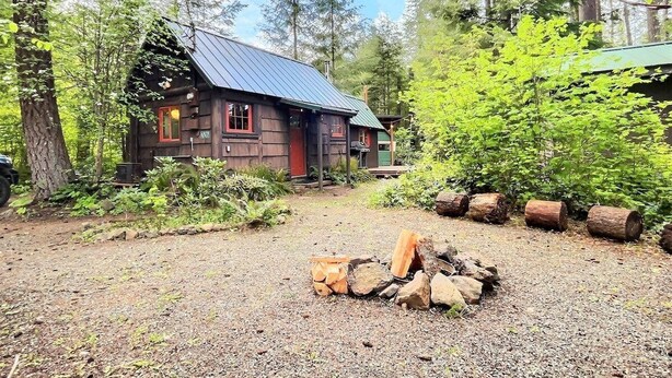 A rustic cabin with a metal roof, surrounded by dense forest. A fire pit with logs is in the foreground. - Ohana Cabin | WiFi, Wood Stove, AC, Hot tub (Packwood)