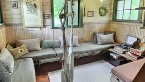 A cozy cabin interior with a wooden ceiling, a hanging swing, a sofa with cushions, a small table with books, and a window with a view. - Ohana Cabin | WiFi, Wood Stove, AC, Hot tub (Packwood)