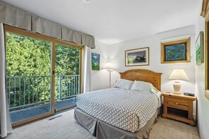 A bedroom with a bed, a nightstand, a lamp, a window with a view of greenery, and framed pictures on the wall. - Central Fish Creek - Hilltop Hideaway Condo (Fish Creek)