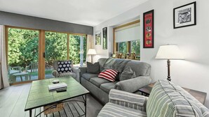 Un salon comprenant un canapé, des fauteuils, une table basse et une lampe. Les murs sont ornés de tableaux encadrés, et une grande fenêtre offre une vue sur les arbres.