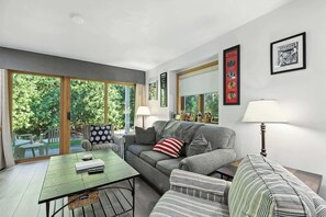 A living room with a sofa, armchairs, a coffee table, and a lamp. There are framed pictures on the wall and a large window with a view of trees.