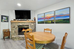 A dining area with a stone fireplace, a round wooden table, and three paintings above the fireplace. - Central Fish Creek - Hilltop Hideaway Condo (Fish Creek)