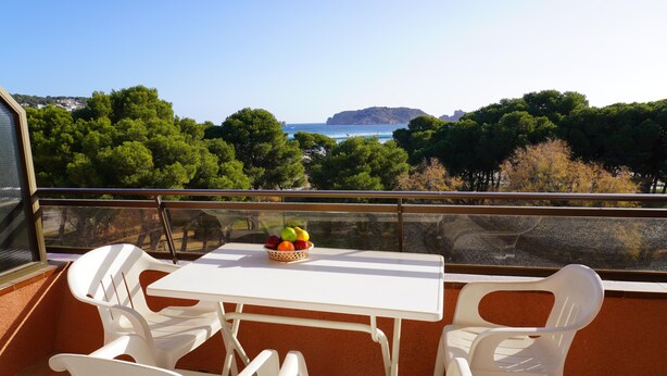 Un balcon avec des chaises en plastique blanches et une table, un panier de fruits sur la table, et une vue sur la mer et les arbres.