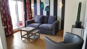 Un salon avec un canapé gris, une petite table, une chaise, un cactus et une porte-fenêtre coulissante.