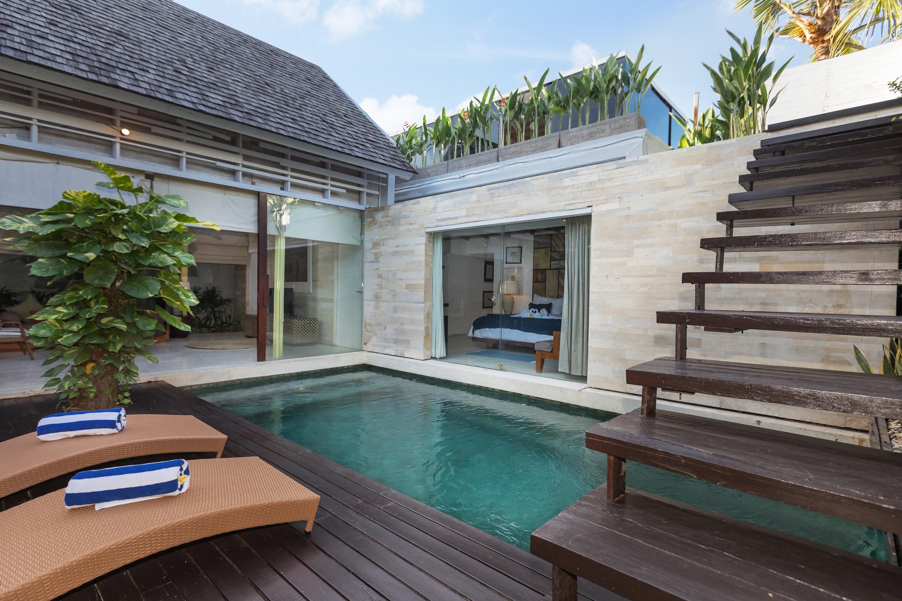 A modern outdoor pool area with wooden loungers and a glass door leading to a bedroom.