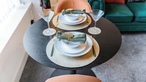 A dining table set with plates, glasses, and cutlery, with a green sofa and colorful cushions in the background. - Cosy Central Aberdeen 1 Bed Flat by Retail Park (Aberdeen)