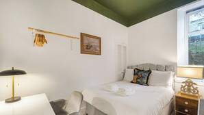 A hotel room with a bed, a desk, a chair, a nightstand, a lamp, a picture on the wall, and a window. - Cosy Central Aberdeen 1 Bed Flat by Retail Park (Aberdeen)
