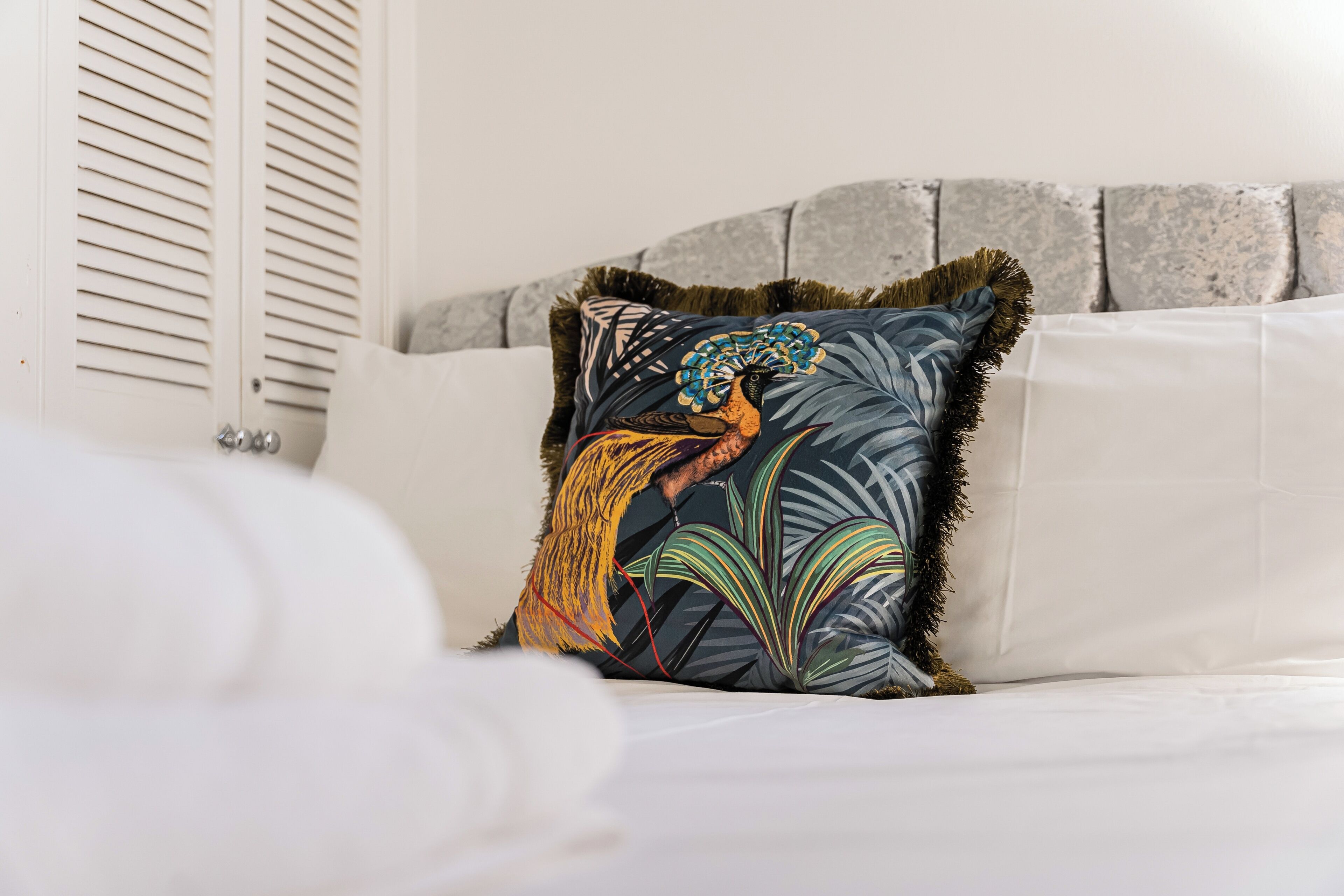 Een bed met een wit hoofdeinde, een decoratief kussen met een tropisch dessin en een witte deken.