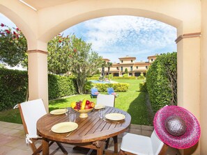 Eine Terrasse im Freien mit einem runden Holztisch, der für ein Essen gedeckt ist, umgeben von weißen Stühlen und einem großen Strohhut auf dem Tisch.