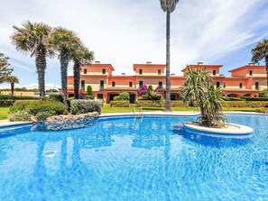 Ein Swimmingpool, umgeben von Palmen und ein mehrstöckiges Gebäude mit einem Ziegeldach.