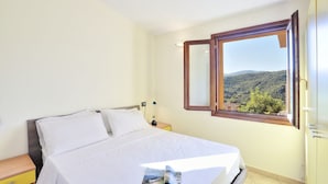 Ein Schlafzimmer mit Bett, Nachttisch, Lampe und einem Fenster mit Blick auf eine hügelige Landschaft.