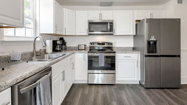 A modern kitchen with white cabinets, stainless steel appliances, a granite countertop, and a window providing natural light. - Comfortable 3 - Bedroom Norfolk Stay  – Minutes from Bases and ODU (Norfolk)