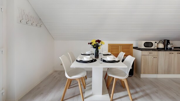 Un coin repas avec une table blanche dressée pour quatre personnes, un vase rempli de fleurs et un espace cuisine équipé d’un four à micro-ondes et de placards.