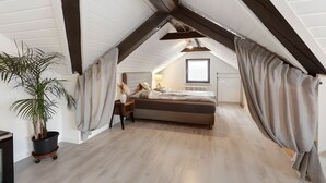 Un dormitorio con techo inclinado, cama, mesita de noche, lámpara, planta en maceta y una ventana.