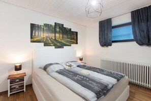 Un dormitorio con cama, mesita de noche, una obra de arte en la pared, ventana con cortinas y lámpara en el techo.