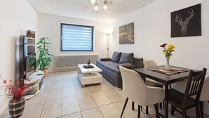 A modern living room with a sofa, coffee table, dining table, chairs, a plant, a radiator, a wall-mounted artwork, and a ceiling light fixture.