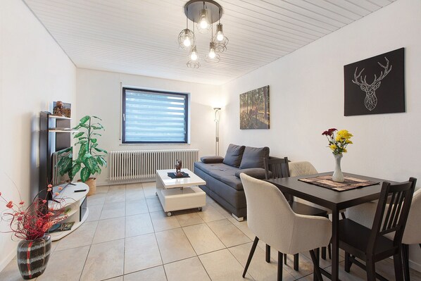 Un salon moderne comprenant un canapé, une table basse, une table à manger, des chaises, une plante, un radiateur, une œuvre d’art accrochée au mur et un luminaire au plafond.