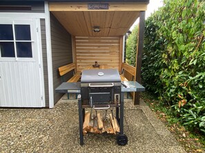 Un espace barbecue extérieur avec un banc en bois et une table, le tout entouré d’une haie.