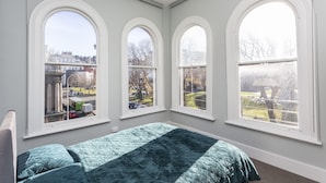 A bedroom with a large bed, three arched windows, and a view of the street outside. - Four bedrooms over looking queens gardens (Dunedin)