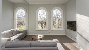 A modern living room with a grey sofa, a round coffee table, and three arched windows offering a view of a park. - Four bedrooms over looking queens gardens (Dunedin)