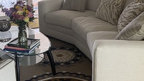 Ein Wohnzimmer mit einem beigefarbenen Sofa, gemusterten Kissen, einem Glastisch mit Büchern, einer Vase mit Blumen und einem Teppich mit Muster.