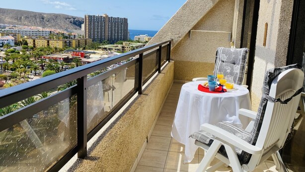 Parvekkeella on aamiasta varten katettu pöytä, ja sieltä avautuu näkymä rannikkokaupunkiin, jossa on rakennuksia ja kauempana vuori.
