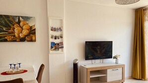 Olohuoneessa on televisio, taulu, valokuvaseinä, ruokapöytä tuoleineen ja pieni kasvi.