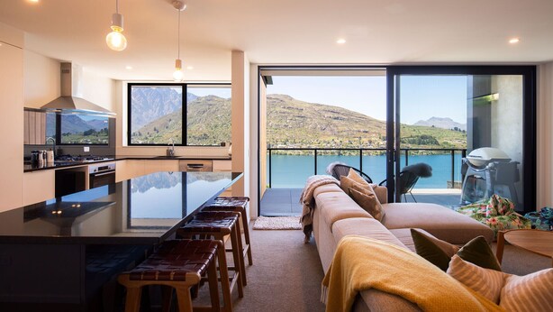 A modern kitchen with a large island, a dining area with bar stools, a sofa with cushions, and a view of a lake and mountains through large windows.