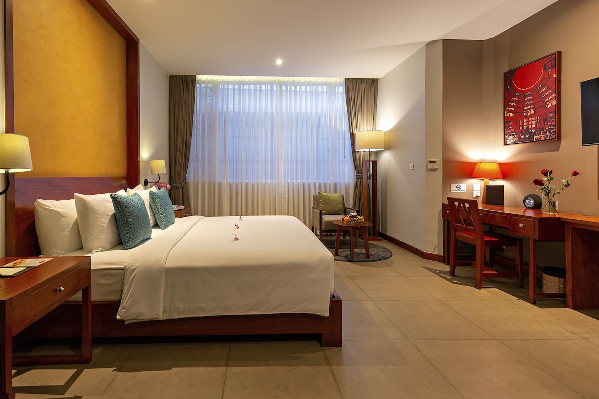 A hotel room with a large bed, two bedside tables with lamps, a desk with a chair, a small table with flowers, and a large window with curtains.