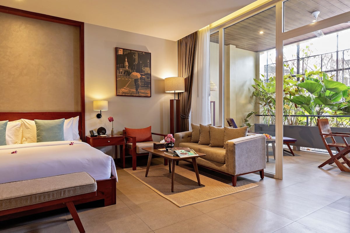 A hotel room with a bed, a sofa, a coffee table, a desk, and a chair. There is a window with curtains and a view of greenery outside.
