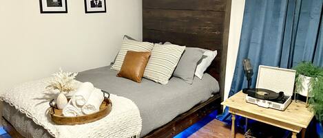 A modern bedroom with a bed, pillows, a bedside table with a record player, and a small plant.