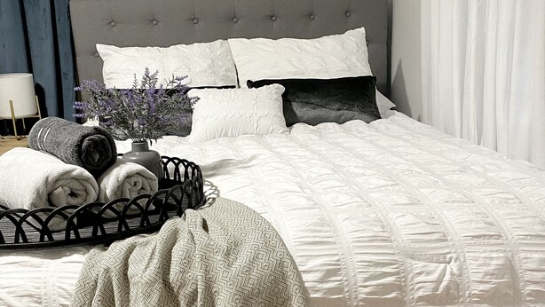 Una cama bien hecha con una colcha blanca, cabecera gris y un cartel decorativo en la pared.