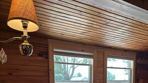 A wooden-paneled room with a round table covered in a floral tablecloth, two windows, and a hanging lamp.