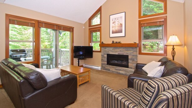 A living room with a fireplace, a television, and a view of the outdoors through large windows. - Upper Benchlands :Great townhouse, corner unit in quiet Treeline. Ski in/ ski out shuttle steps away (Whistler)