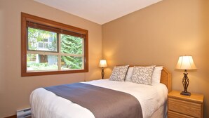 A bedroom with a large bed, two bedside lamps, a window with a view of greenery, and a wooden nightstand. - Upper Benchlands :Great townhouse, corner unit in quiet Treeline. Ski in/ ski out shuttle steps away (Whistler)