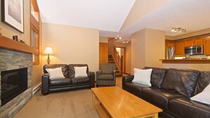 A living room with a fireplace, two leather sofas, a wooden coffee table, and a kitchen area in the background. - Upper Benchlands :Great townhouse, corner unit in quiet Treeline. Ski in/ ski out shuttle steps away (Whistler)
