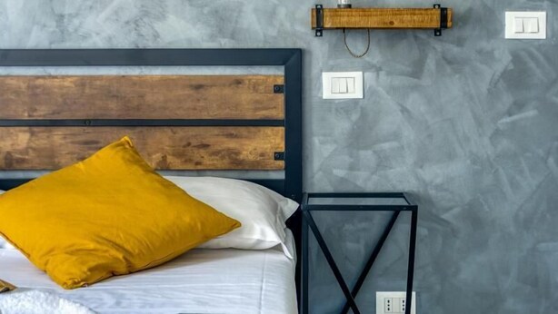 A bedroom with a wooden headboard, a bed with white and yellow bedding, a bedside table with a lamp, and a textured gray wall. - SAPIENZA Grand Apartment (ROMA)