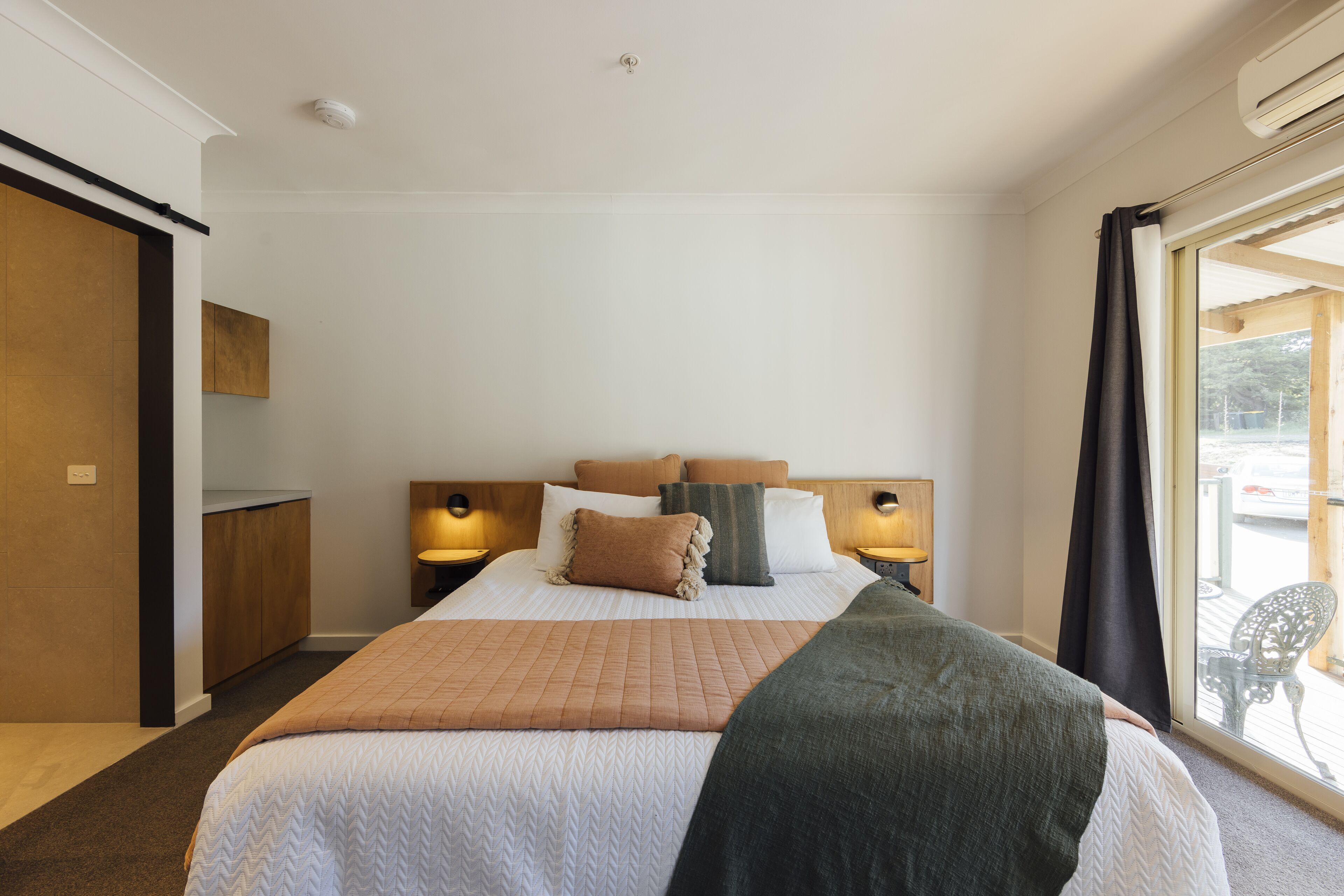 A hotel room with a large bed, two bedside tables with lamps, a wooden headboard, a sliding door, and a view of the outdoors.