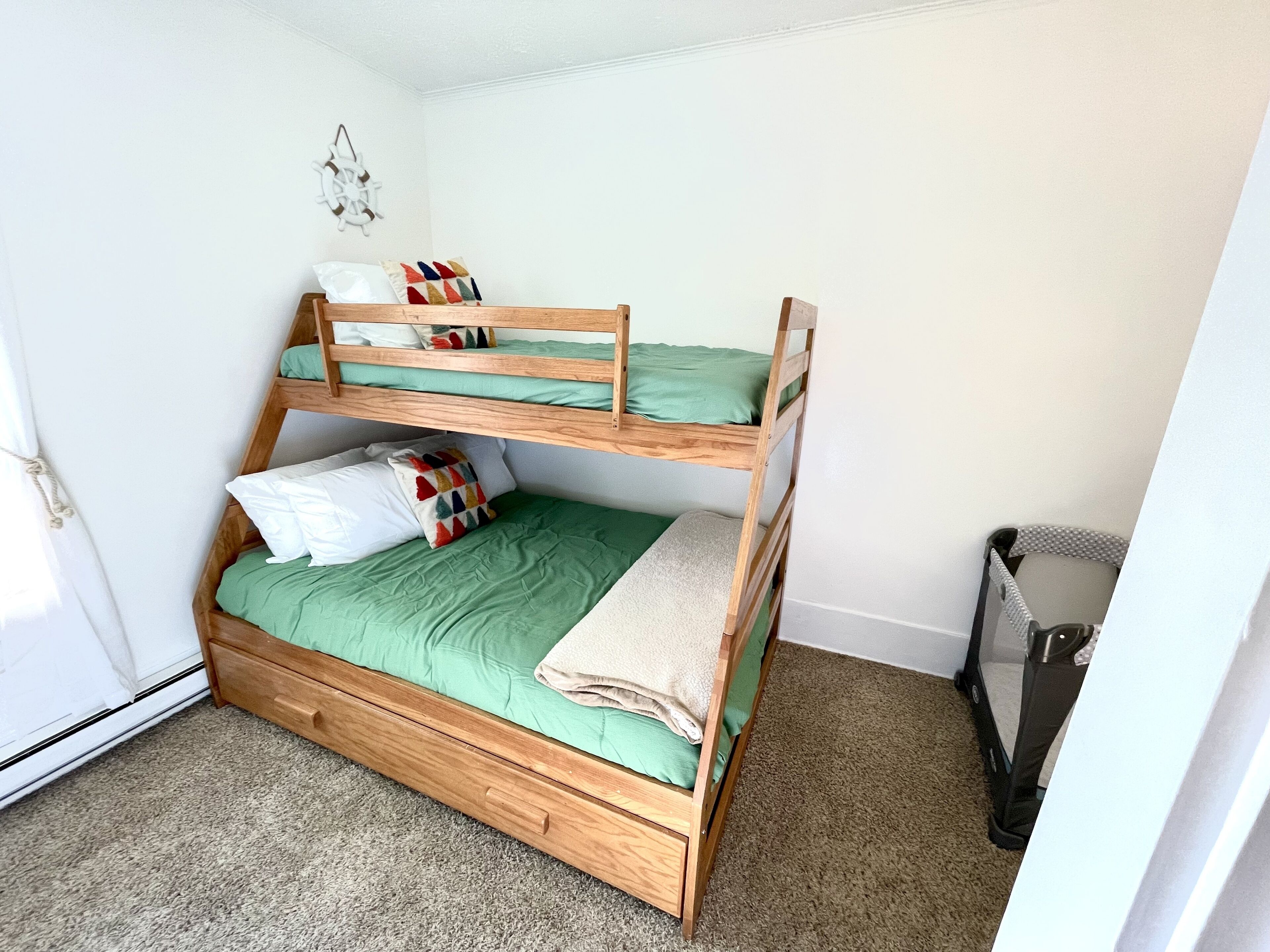 Un letto a castello con biancheria verde e molti cuscini, un piccolo cassetto in legno e un cestino nella stanza, con pareti bianche e pavimento rivestito di moquette.