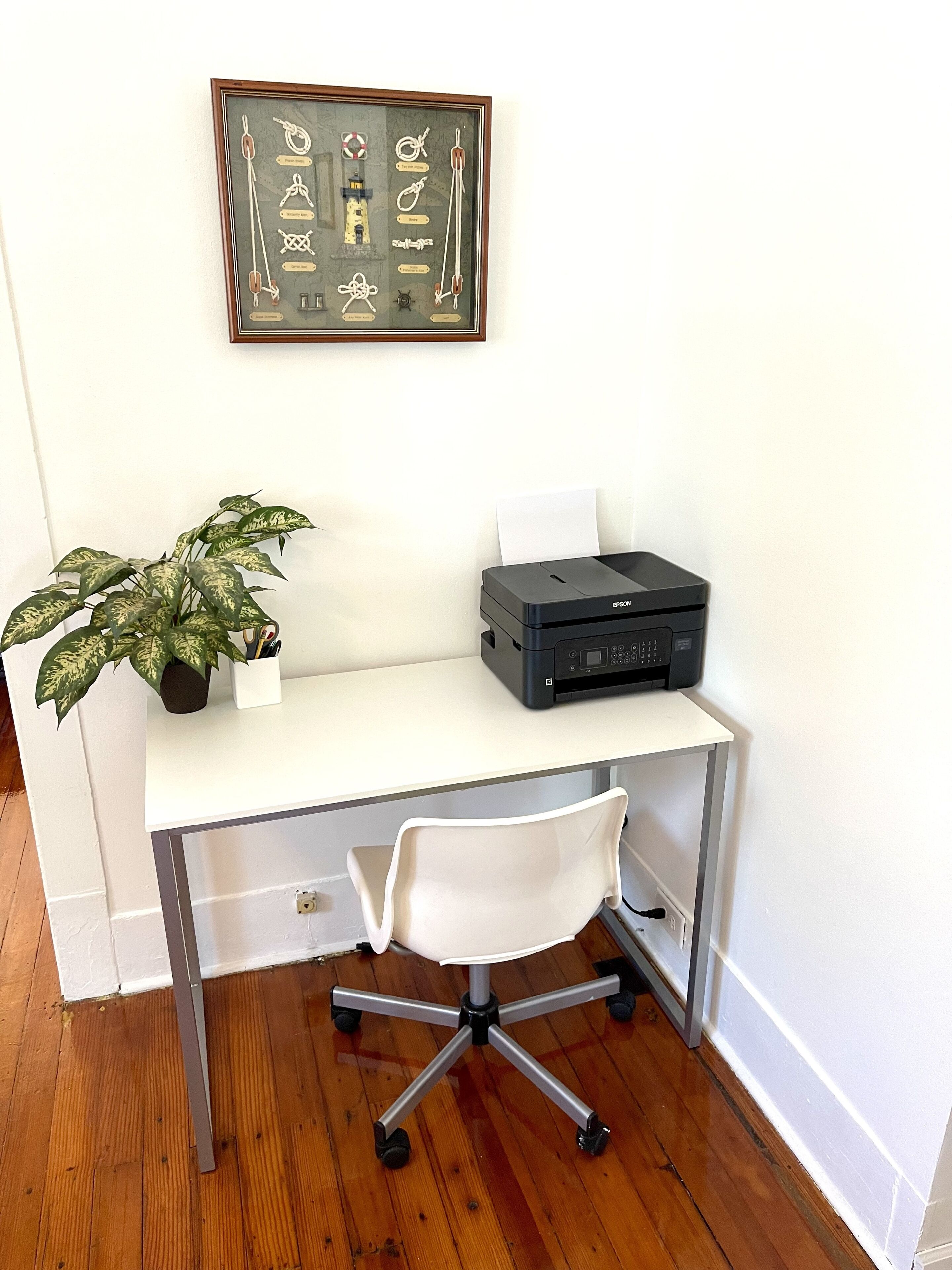 A white desk with a printer, a potted plant, and a framed nautical chart on the wall.